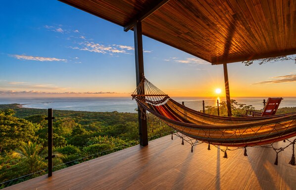 Una hamaca en una terraza de madera con vistas a un paisaje costero al atardecer.