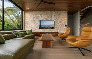 A modern living room with a wooden ceiling, a stone textured wall, a large window, a wooden coffee table, a sofa, and two leather armchairs.