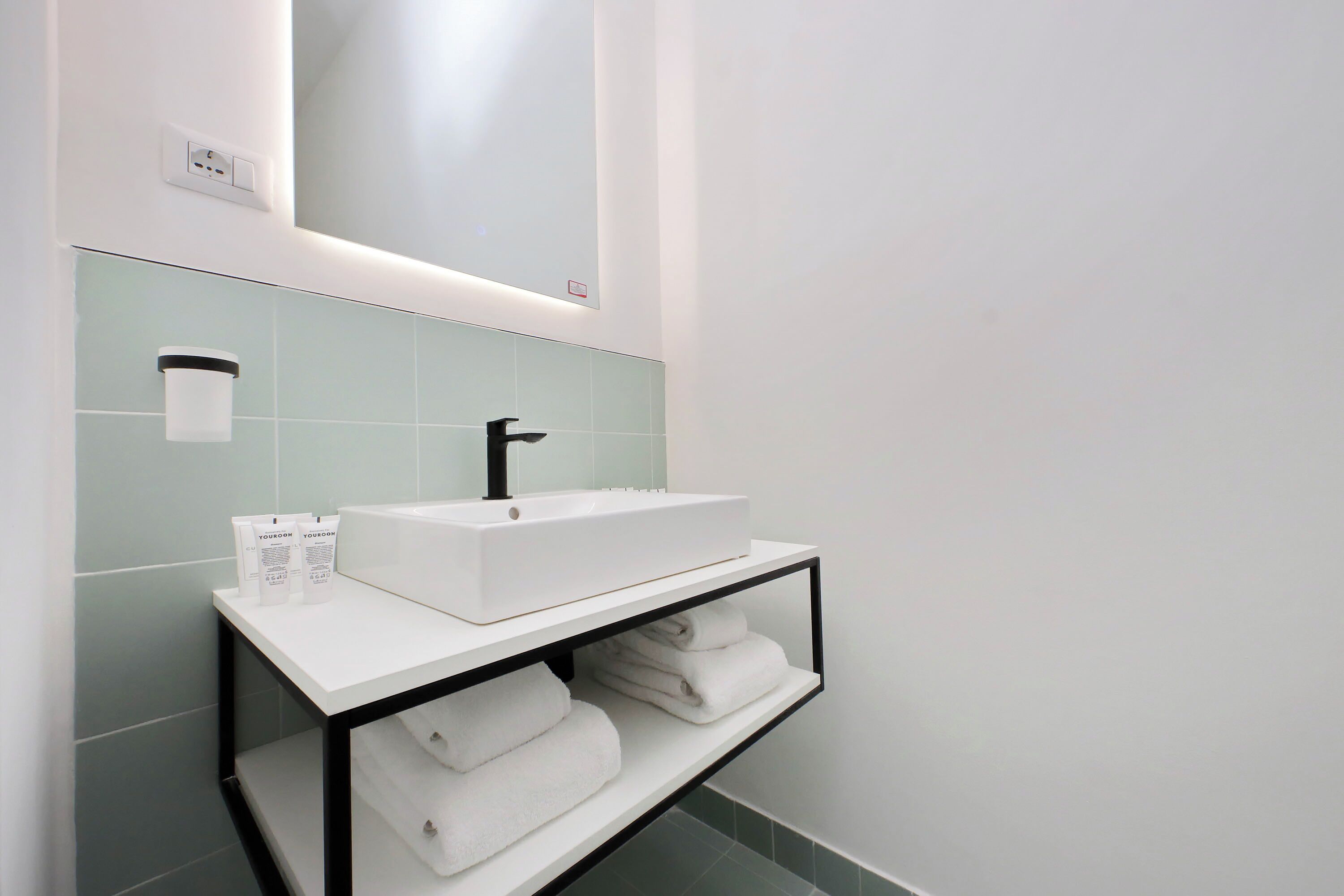 Une salle de bain moderne avec un lavabo blanc, un miroir et une étagère où sont pliées des serviettes.