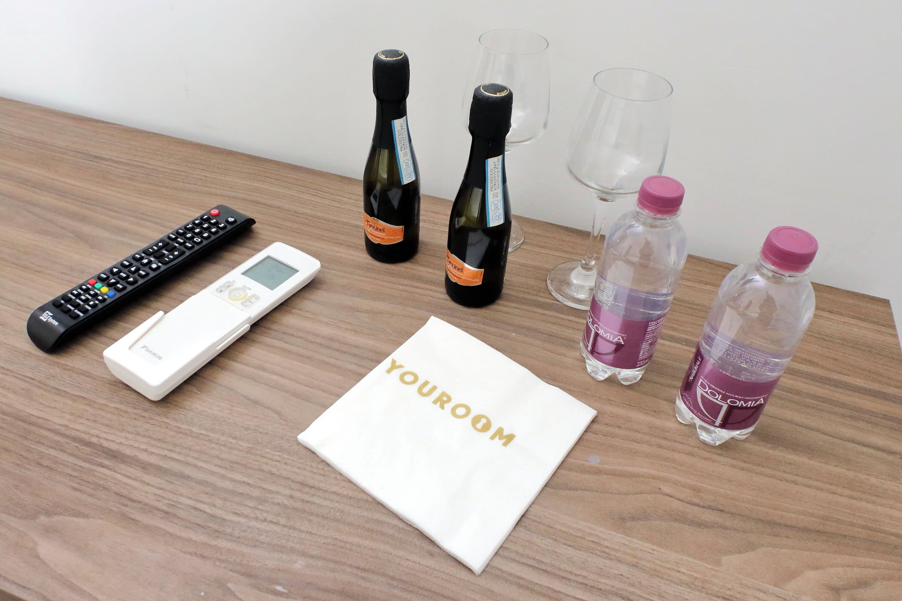 Una mesa de madera con un control remoto, una balanza digital, dos botellas de vino, dos copas de vino vacías, un mantel blanco con el texto "YOROOM" y dos botellas de agua de plástico.