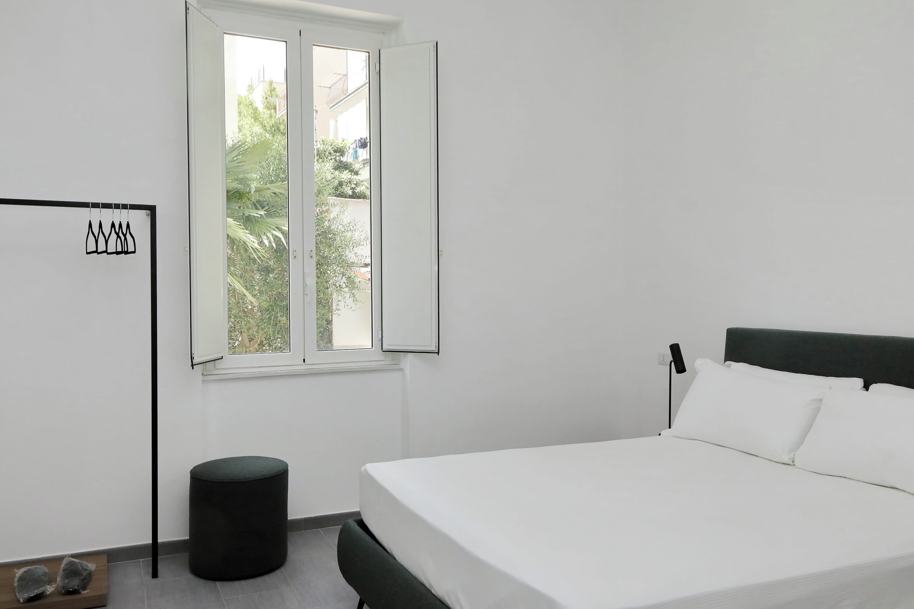 Une chambre avec un lit, une fenêtre, un petit tabouret et une étagère fixée au mur.
