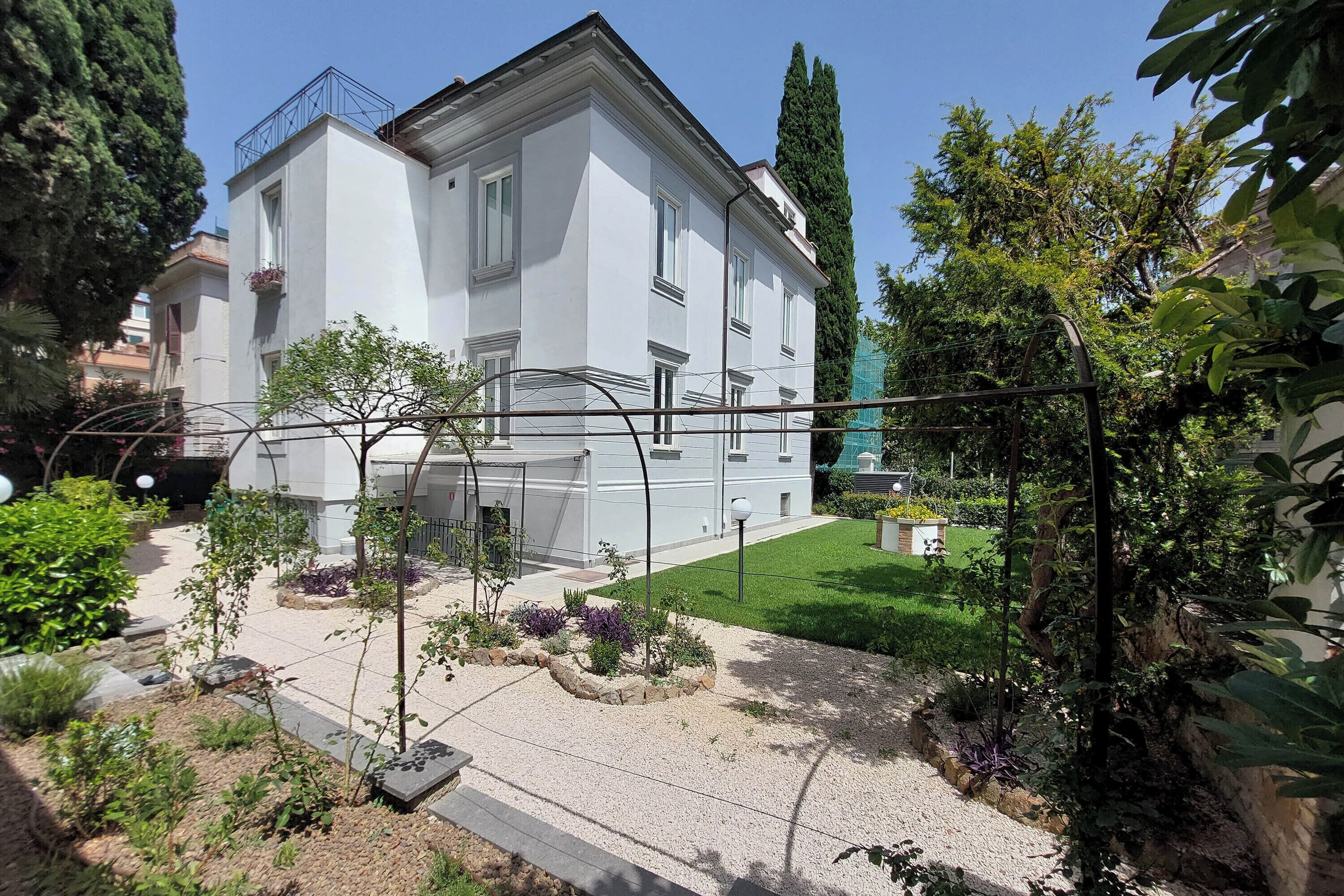 A white building with a balcony, surrounded by a garden with various plants and a gravel path.