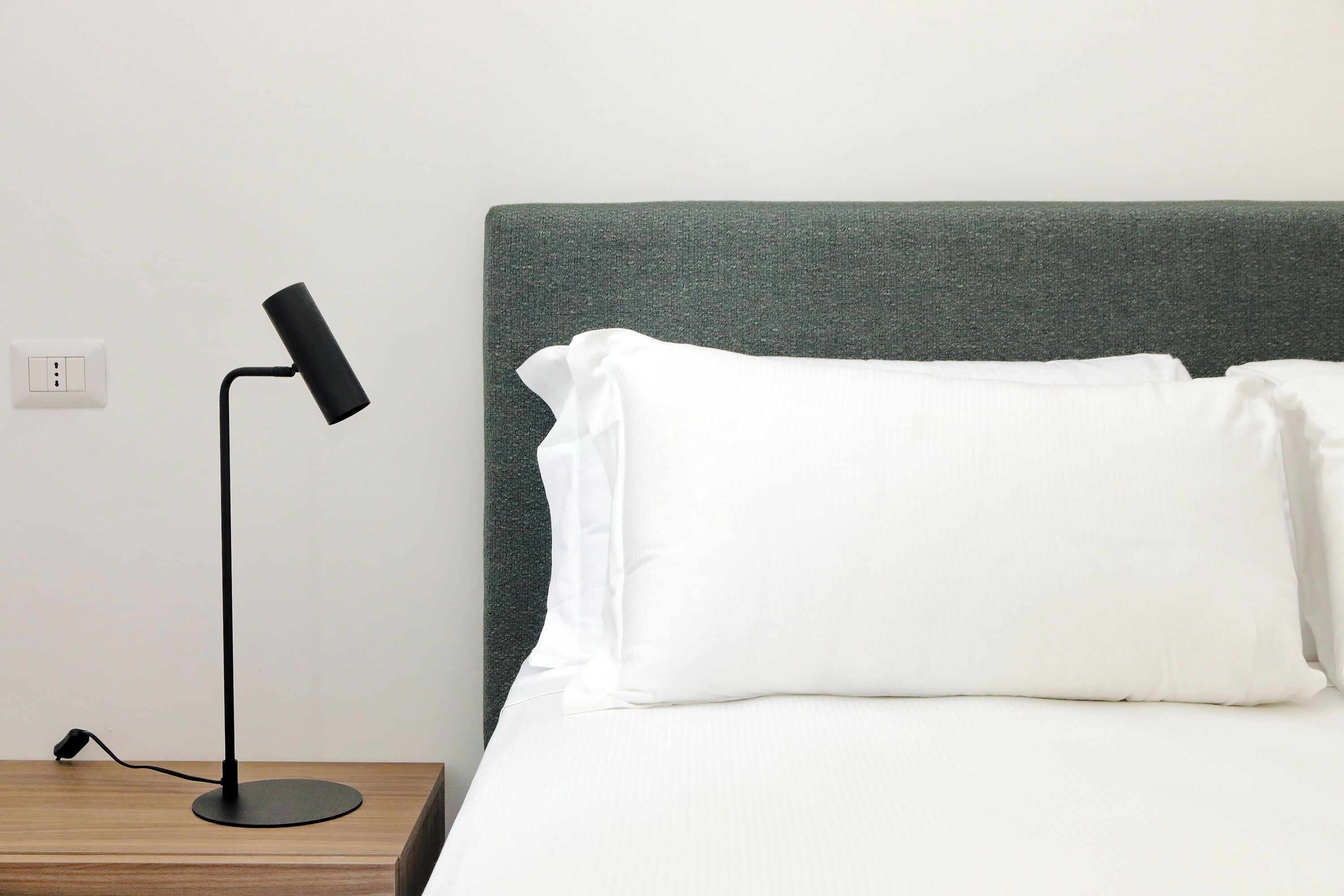 Una cama bien hecha con almohadas blancas y un cabecero verde, una lámpara de escritorio negra y un enchufe en la pared.