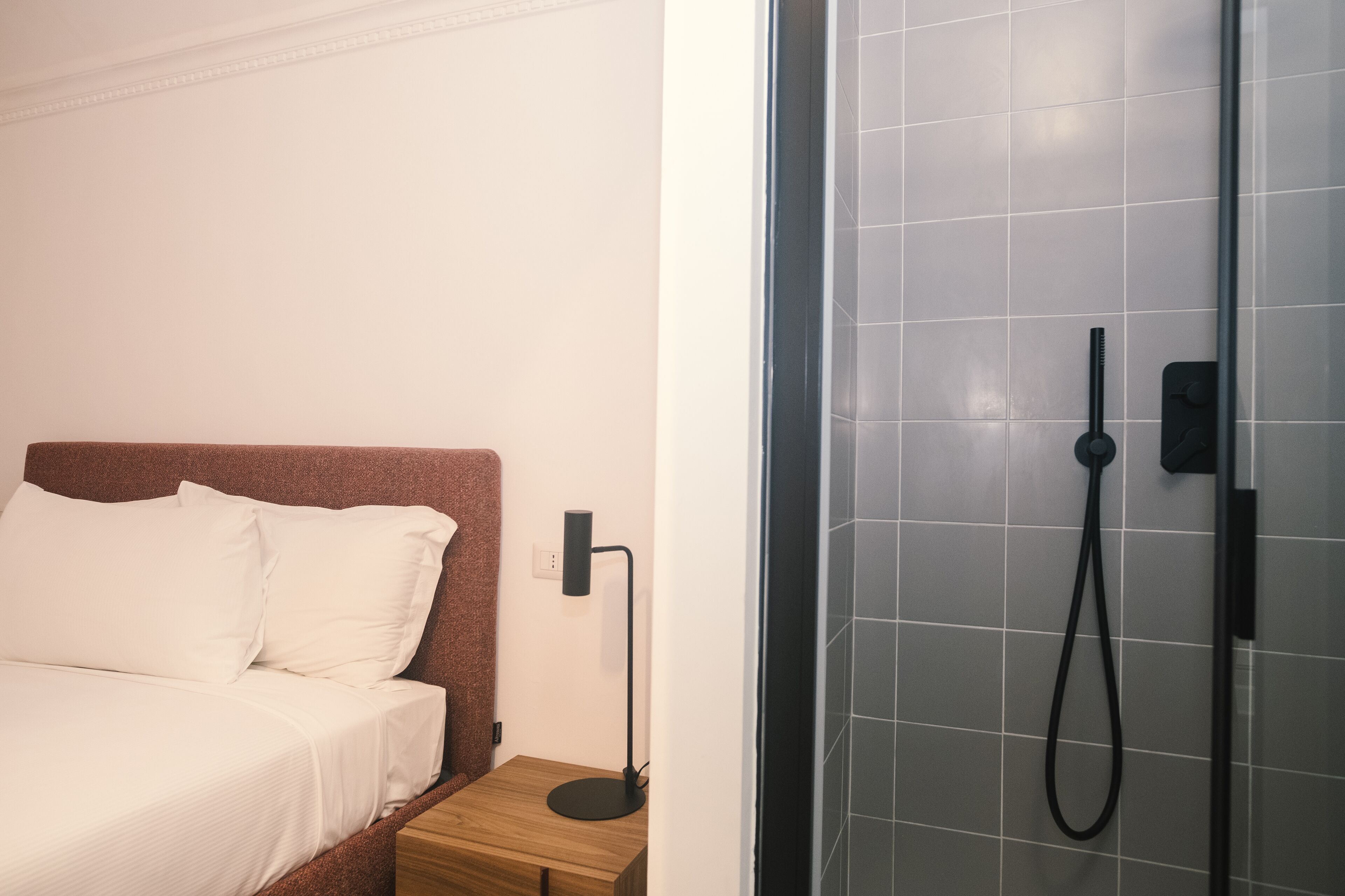 A bedroom with a bed, bedside table, and a modern shower with a handheld showerhead.