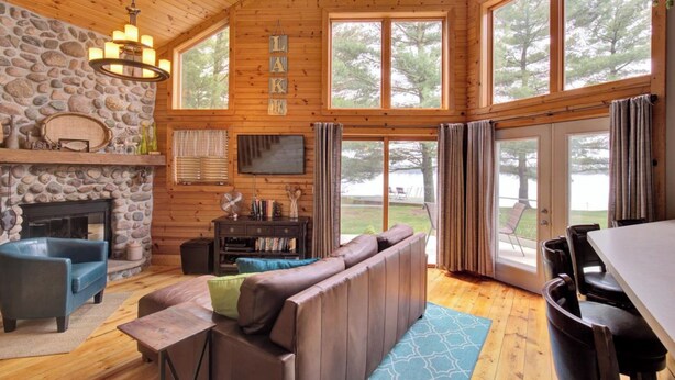 A cozy cabin interior with a stone fireplace, wooden walls, and large windows offering views of the outdoors. - Lake front cottage less than 20 min from golf simulator/pool/bar/grill (Arkdale)