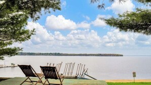 A wooden deck with two lounge chairs facing a calm lake, a pier extending into the water, and a grassy area with a sign. - Lake front cottage less than 20 min from golf simulator/pool/bar/grill (Arkdale)