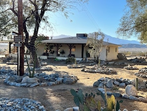 Une maison de style désertique, dotée d’un patio et entourée de rochers et de cactus, avec des montagnes en arrière-plan.