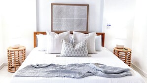 A neatly made bed with a white comforter and gray throw blanket, flanked by two wooden nightstands with lamps, a large abstract wall art piece above the bed, and a ceiling fan above.