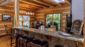 A rustic wooden cabin interior with a stone countertop bar, wooden chairs, a sink, and a chalkboard displaying a Wi-Fi password. - 4-Bed/Bath Cabin: Hot Tub, Game Room, Pet-Friendly! (Seven Devils)