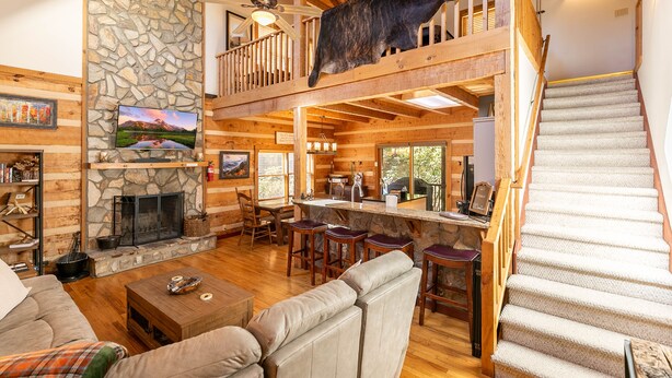 A spacious living area with a stone fireplace, wooden ceiling beams, a large bear mounted on the wall, and a staircase leading to an upper level. - 4-Bed/Bath Cabin: Hot Tub, Game Room, Pet-Friendly! (Seven Devils)
