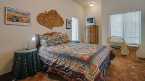 A bedroom with a bed, a nightstand, a lamp, a wicker chair, a cabinet, and a window with blinds.