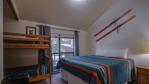 A bedroom with a bunk bed, a large bed with a colorful blanket, a window with curtains, and skis hanging on the wall. - Comfy Condo near Shuttle Pick up (Angel Fire)