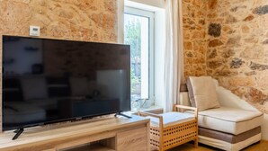 A living room with a flat-screen TV on a wooden stand, a stone wall, a window with curtains, and a sofa. - Thrapsano House for up to 8 Persons, Near Iraklion (Minoa Pediada)