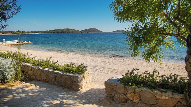 A beach with clear blue water, a boat on the shore, and a stone wall. - Vanellus Villas Sleeps 6 With Jacuzzi (Pakostane)