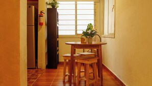 A small kitchen with a refrigerator, a window with blinds, a wooden table with two stools, and a potted plant on the windowsill. - Loft in Roma Sur: King Size Bed (Cuauhtémoc)