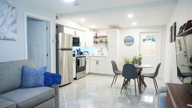 A modern kitchen with stainless steel appliances, a dining area with a round table and chairs, and a living room with a sofa and a blue pillow. - Cozy 1 bedroom suite-apartment. (Port Richey)
