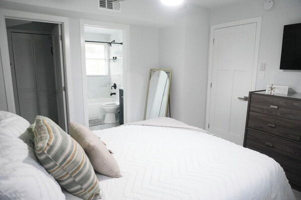 Un dormitorio con cama, cómoda, espejo y un baño visible a través de una puerta abierta.