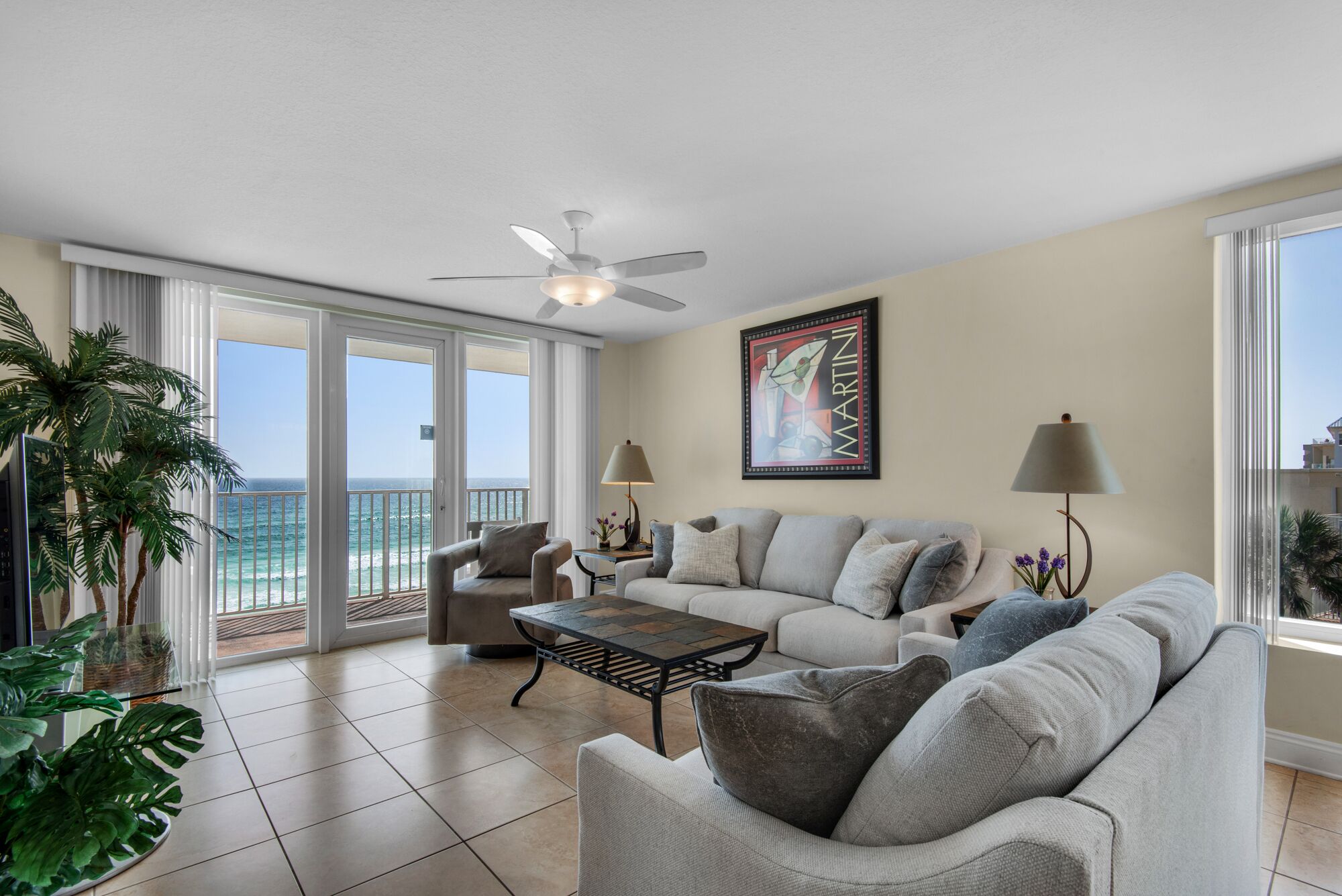 A spacious living room with a large gray sofa, a coffee table, and a ceiling fan. There is a large window with a view of the ocean.