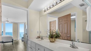 Un baño de hotel con un espejo grande, dos lavamanos, una planta en maceta y vistas al océano a través de la habitación contigua.