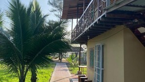 Un sendero lleva a un edificio con balcón cubierto, palmeras y una zona con pavimento de ladrillo.