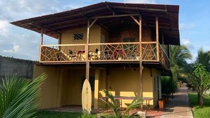 Un edificio de dos pisos con un balcón, una tabla de surf y un camino que lleva a la entrada.