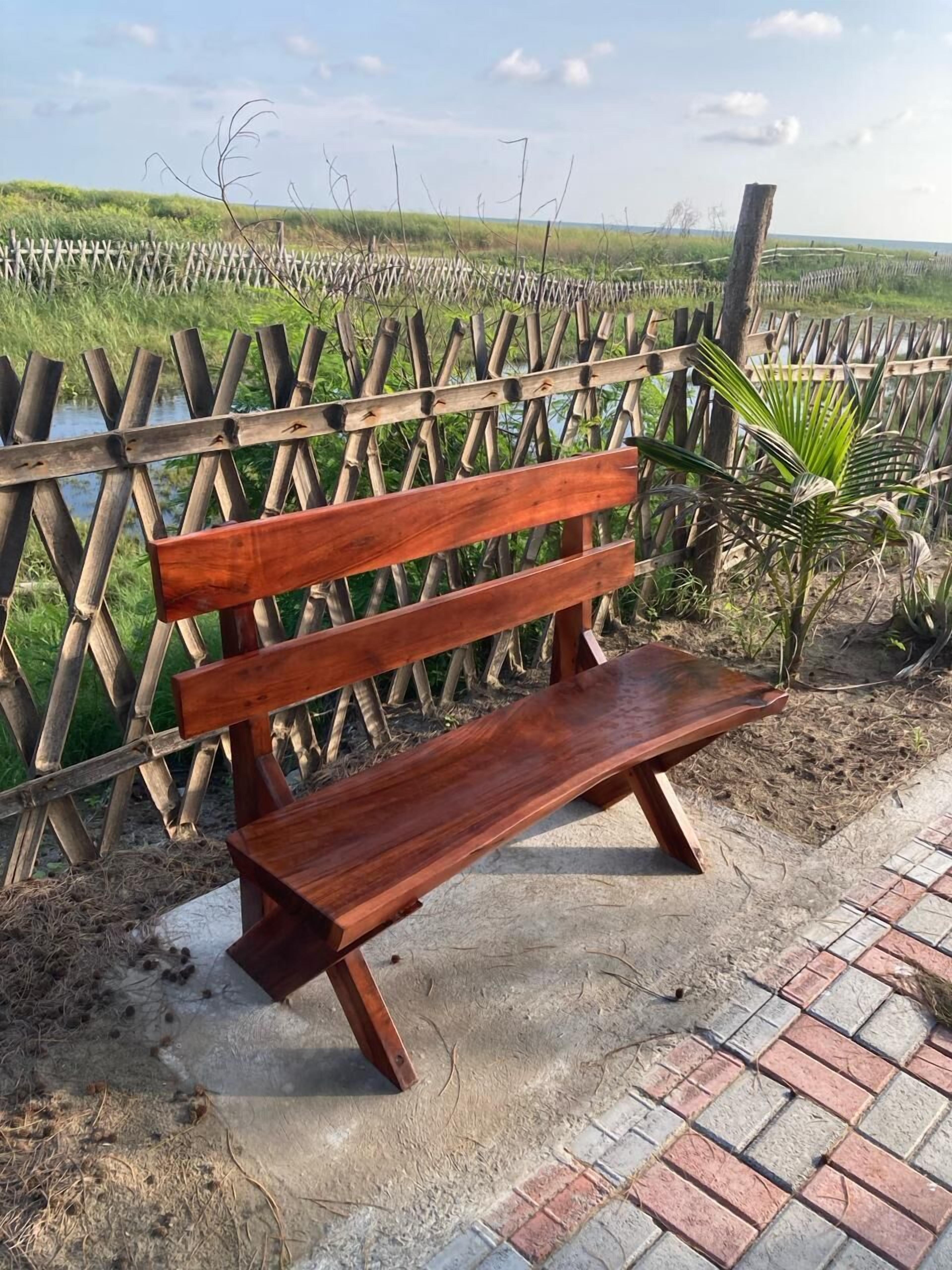 Un banc en bois doté de pieds en métal, placé sur une zone pavée avec un motif en briques, à côté d’une clôture en bois, avec un paysage naturel en arrière-plan.