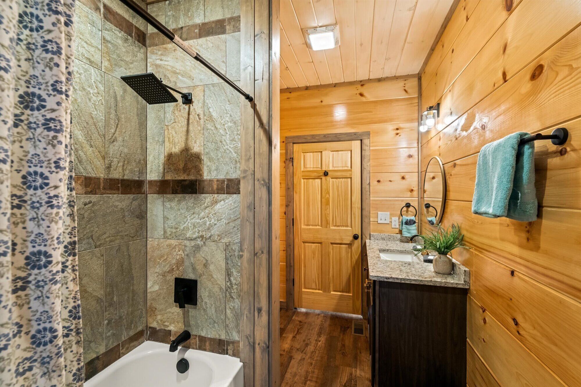 Un baño con ducha de piedra, paredes de madera, una puerta de madera, un lavabo con espejo y una toalla colgada en un gancho.