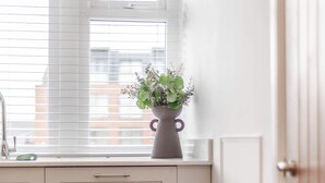Een keuken met witte kastjes, een raam met jaloezieën, een vaas met bloemen en een ronde tafel met stoelen.