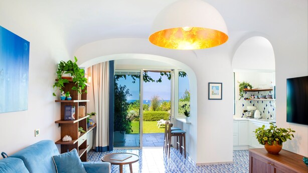 A modern living room with a blue sofa, a coffee table, and a bookshelf. There is a large window with a view of the outdoors. - PRESTIGE SEA VIEW VILLA SCANNELLA (Forio)