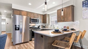 A modern kitchen with stainless steel appliances, a central island with bar stools, wooden cabinets, and a hanging pendant light. - Brand New Dog-Friendly Getaway! Perfectly Positioned for Boise Adventures! Walk to Grocery & More! (Boise)