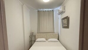 Een kleine kamer met een enkel bed, houten vloer, witte muren en een plafondlamp.