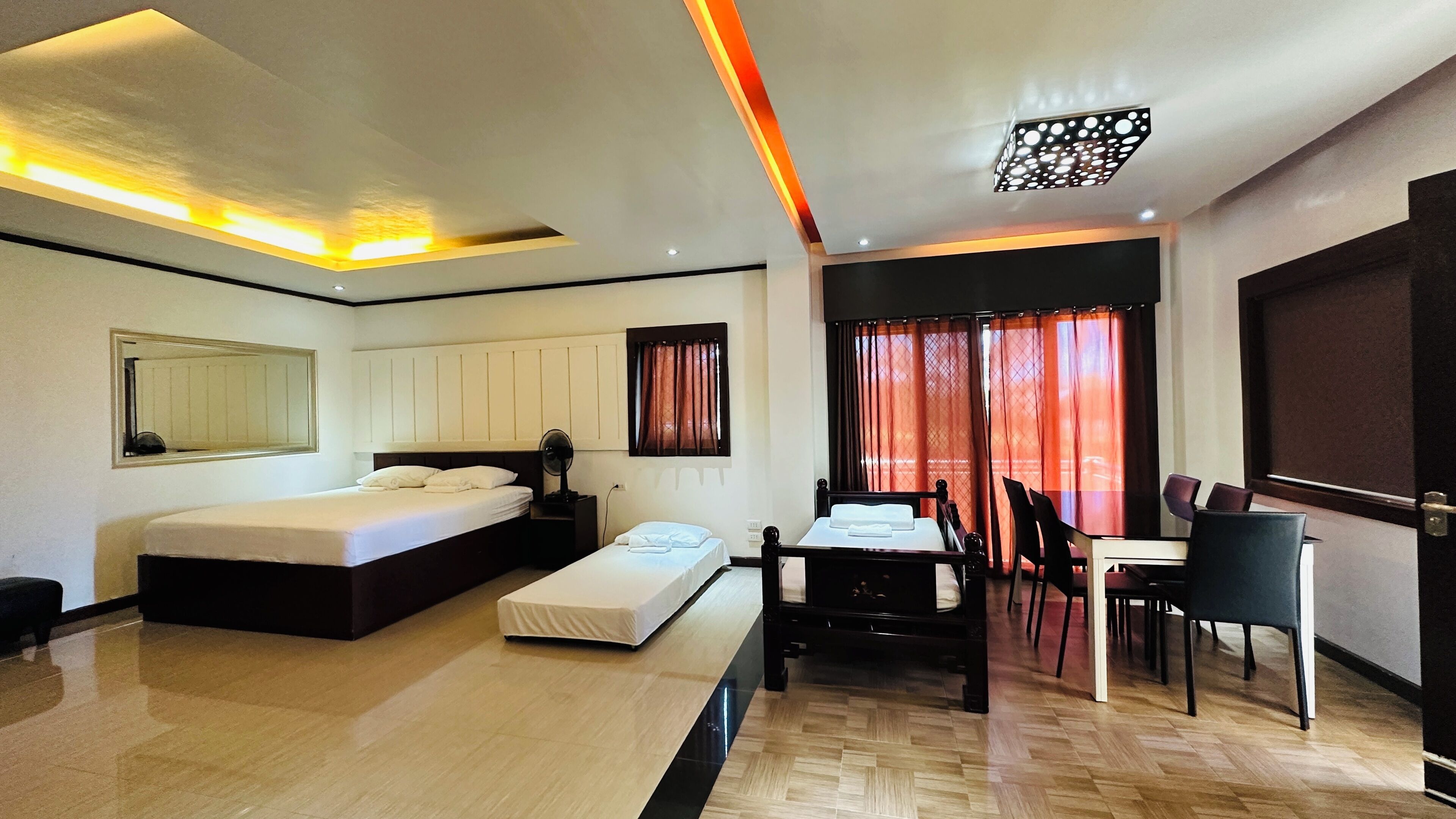 A modern hotel room with a large bed, a desk with chairs, a dining table, and a view of the outside through red curtains.