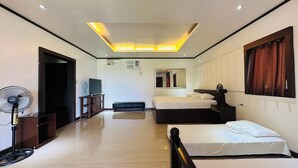 A hotel room with a bed, bedside table, a desk with a chair, a standing fan, a TV, and a ceiling light fixture. - Tugsaw Resort (El Salvador)