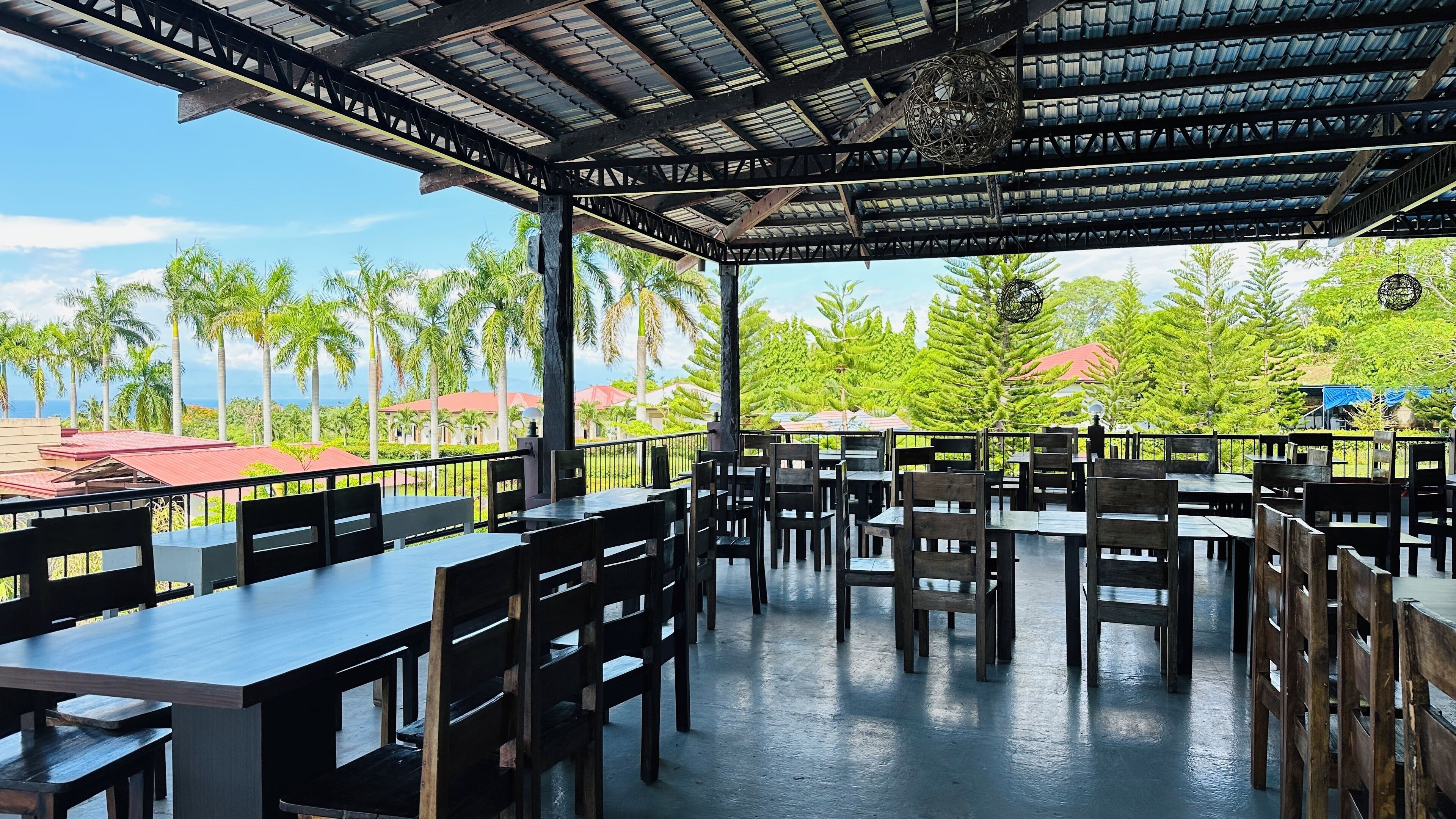 An outdoor restaurant with tables and chairs, a covered roof, and a view of palm trees and buildings.