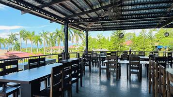 An outdoor restaurant with tables and chairs, a covered roof, and a view of palm trees and buildings.