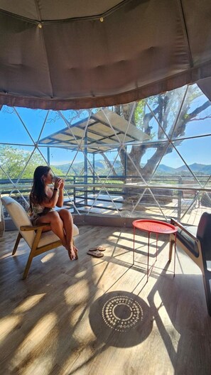 Uma pessoa sentada numa cadeira sob um toldo, com vista para árvores e montanhas através de uma estrutura de vidro.