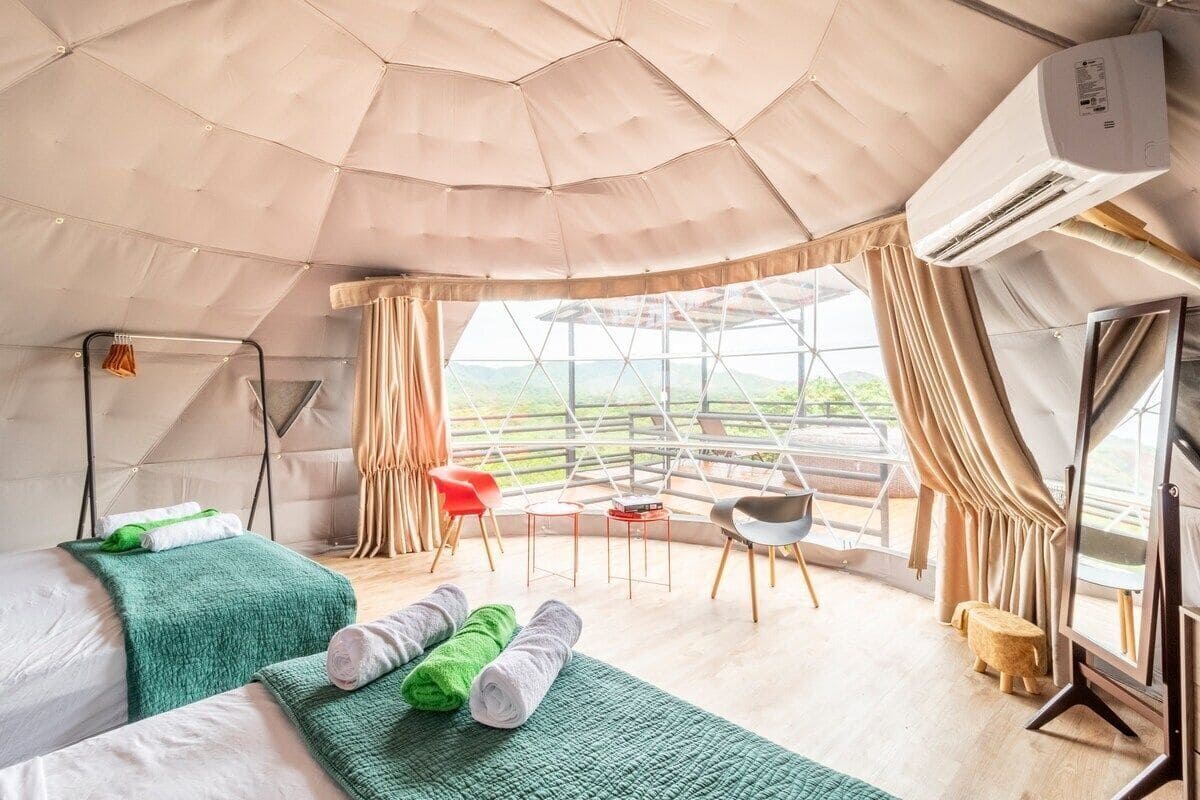 A tent-style room with a bed, green towels, a small table, a chair, and a window with curtains.