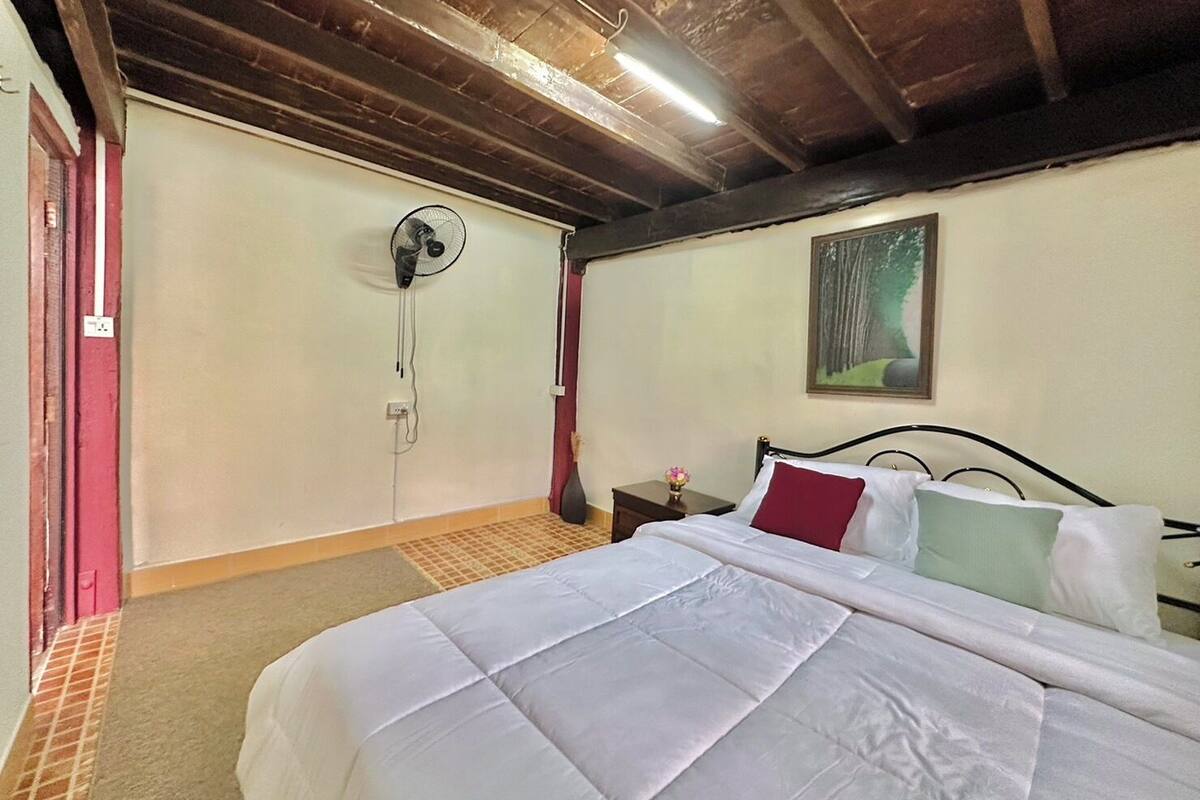 Une chambre à coucher avec un lit, un ventilateur fixé au mur, une petite table de chevet avec un vase et un tableau accroché au mur.