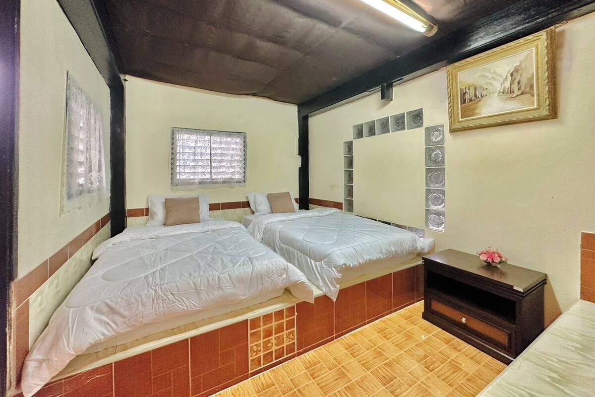 Une chambre avec deux lits, une fenêtre avec des stores, un miroir orné de motifs décoratifs et une petite table sur laquelle est posée une fleur.