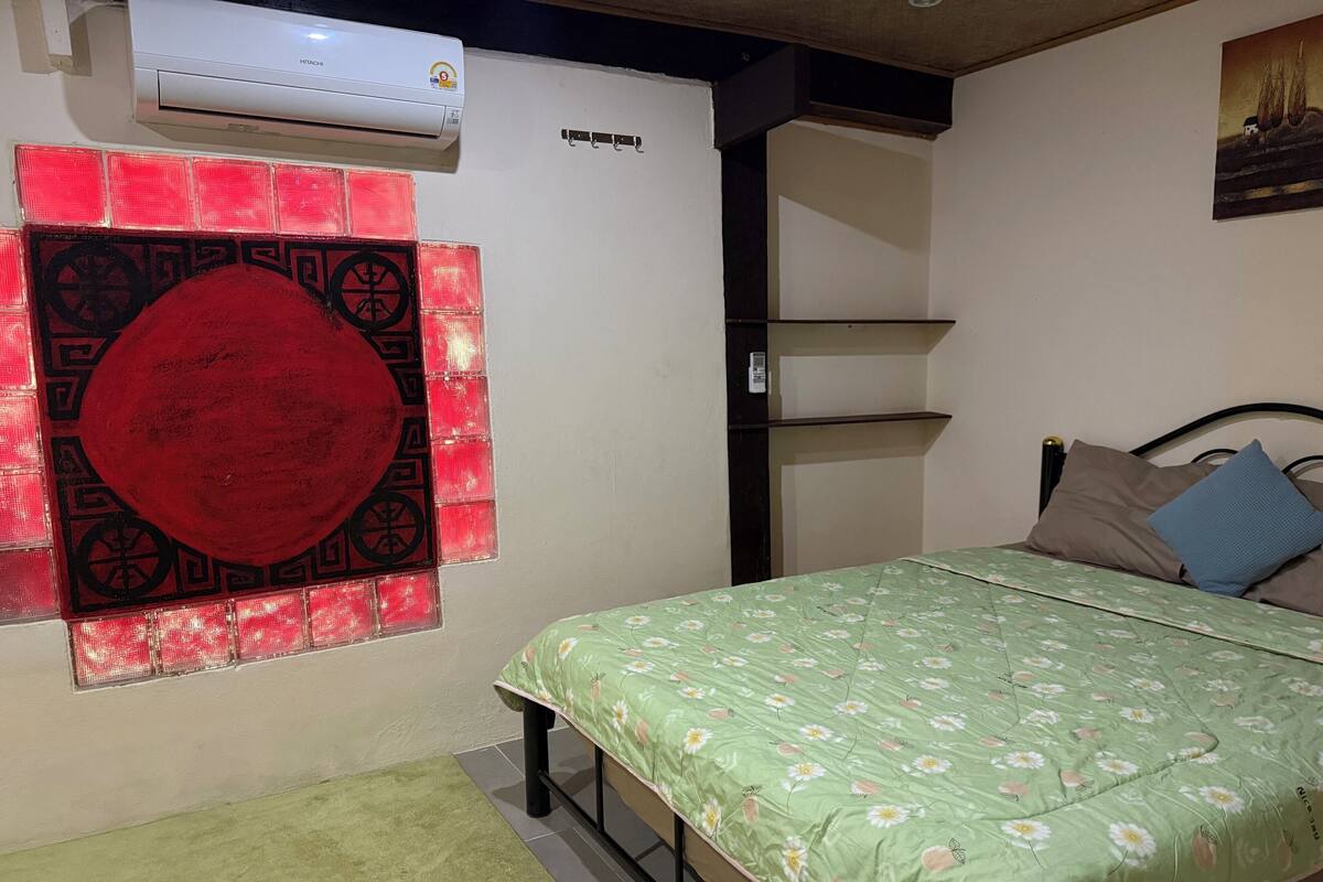 Une chambre à coucher avec un lit, un élément décoratif rouge au mur, une moquette verte et un appareil de climatisation.