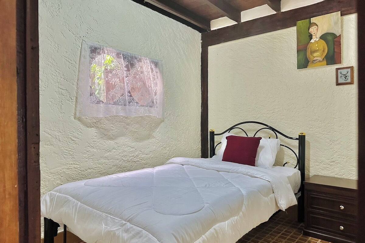 Une chambre à coucher avec un lit, une table de chevet, une tête de lit en bois, un mur orné d’un tableau et une fenêtre avec un rideau en dentelle.