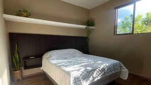 Een slaapkamer met een bed, een plafondlamp, een raam met uitzicht op groen en een plank met planten.
