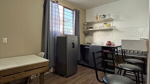 Een kleine keuken met een koelkast, een spoelbak en een fornuis. Er is een eettafel met stoelen en een raam met gordijnen.