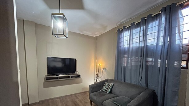 Een woonkamer met een flatscreen televisie aan de muur, een grijze bank met een gekleurd kussen, een hangende hanglamp en een vloerlamp.