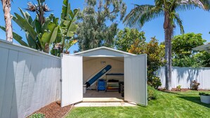 A small, open garage with a blue kayak and a white shed in a backyard with a lawn and various plants. - Leucadia "Ponto" Bungalow ~ walk everywhere with beach gear! (Encinitas)