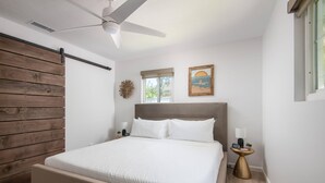 A bedroom with a bed, a ceiling fan, a wooden wall panel, a window, and a small table with a lamp. - Leucadia "Ponto" Bungalow ~ walk everywhere with beach gear! (Encinitas)