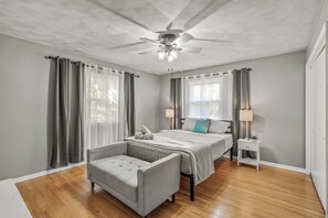 A bedroom with a bed, two bedside tables, a chair, a ceiling fan, and a window with curtains.