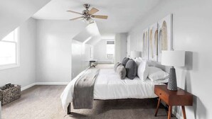 Quarto com cama, mesa de cabeceira, ventilador de teto e janela.
