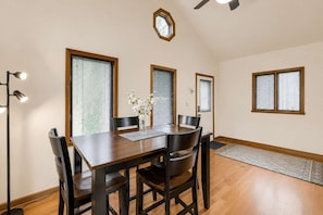 A dining area with a wooden table, chairs, and a vase of flowers. There are windows with blinds, a ceiling fan, and a rug on the floor.
