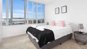 Une chambre moderne avec une grande fenêtre, un lit agrémenté d’oreillers blancs et roses, un plaid noir et une table de chevet avec une lampe.